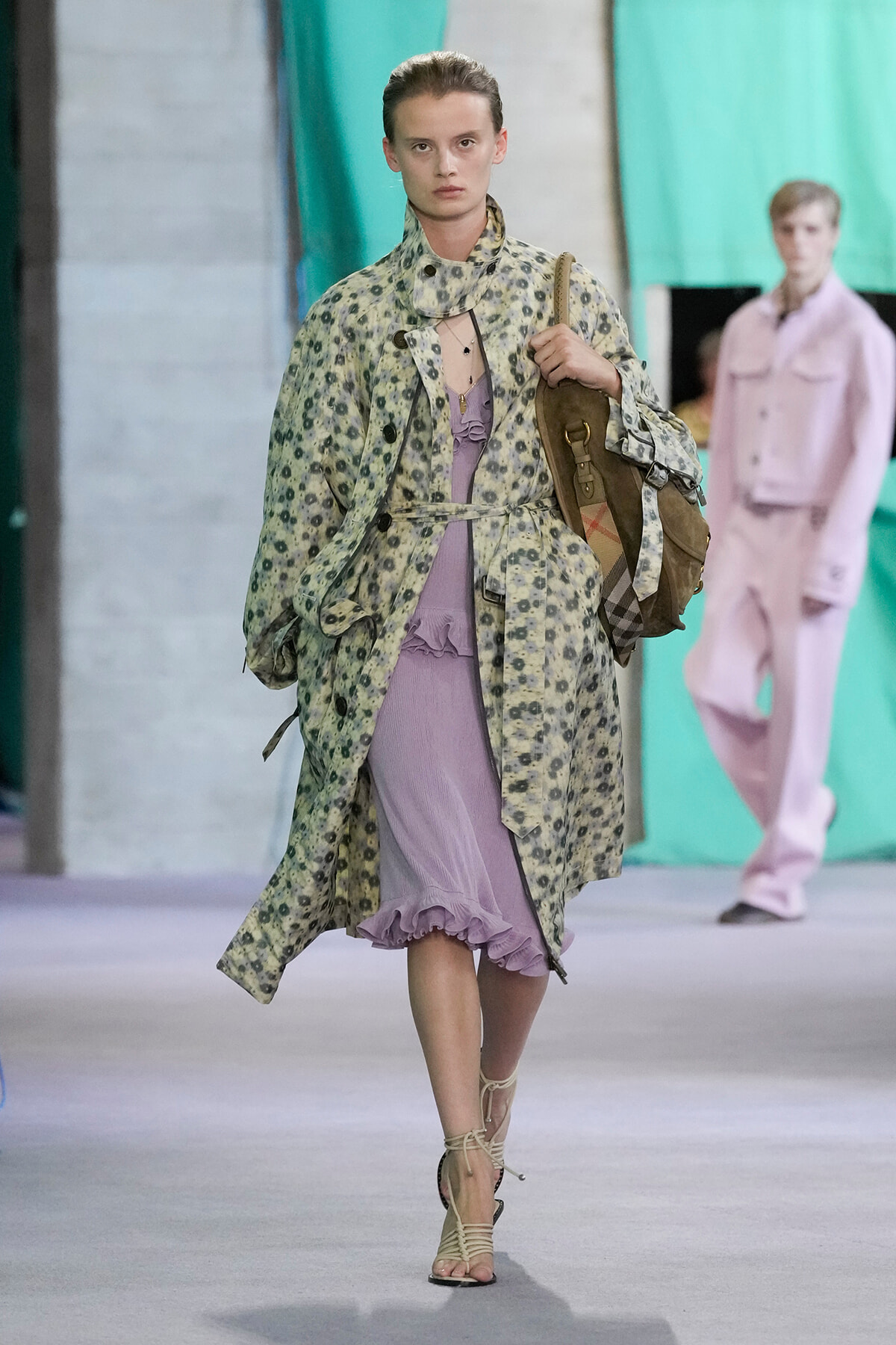 Female runway model walking in a pale floral trench over a lavender ruffled dress, carrying a brown satchel and wearing strappy sandals.