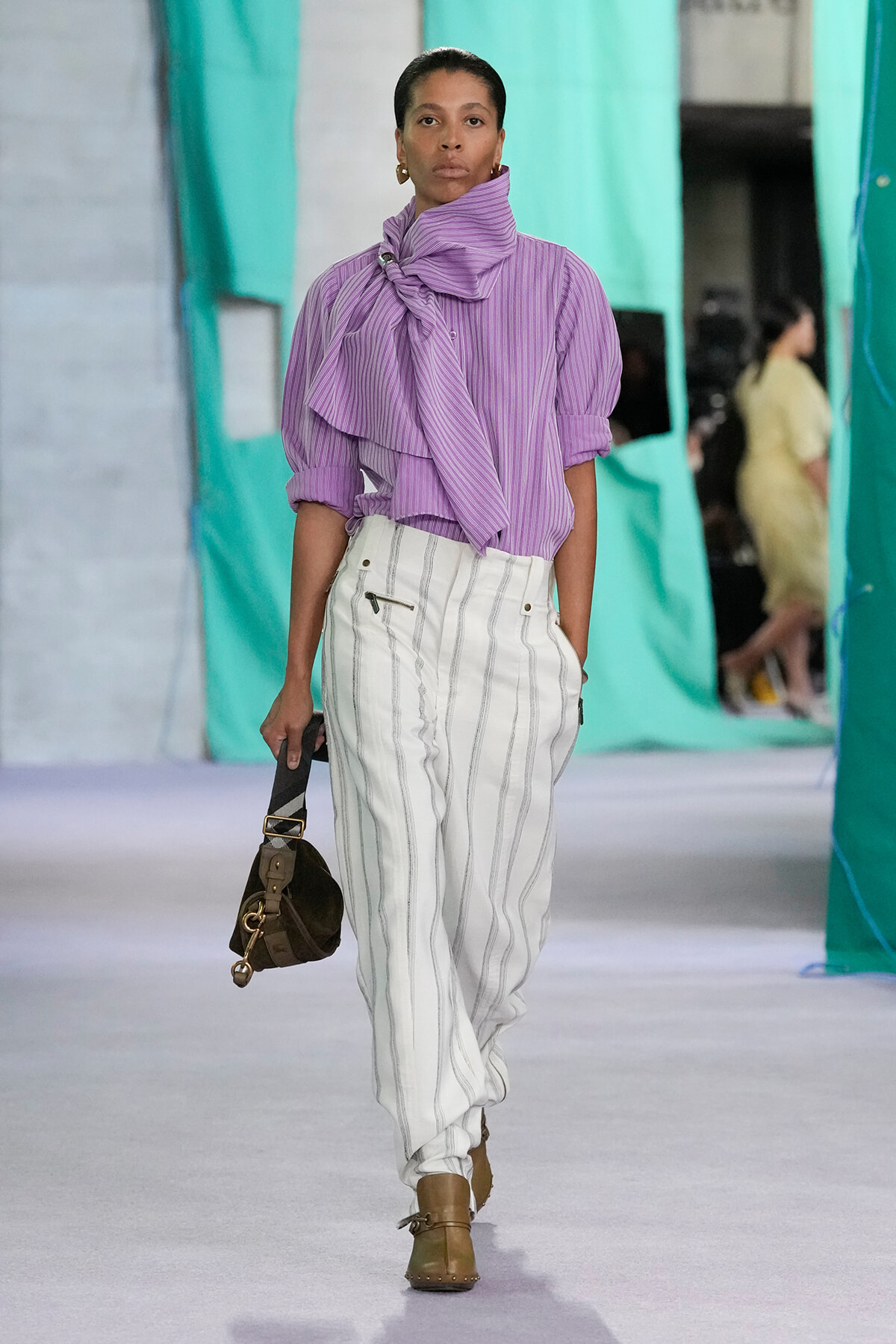 Model walking a fashion show wearing a lavender striped blouse with a large neck bow, white pinstriped trousers, and tan ankle boots carrying a brown bag.
