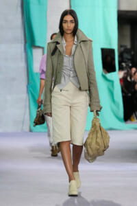 Female model walks a fashion runway in a light gray-green jacket over a striped shirt and beige shorts, carrying a large tan quilted bag.