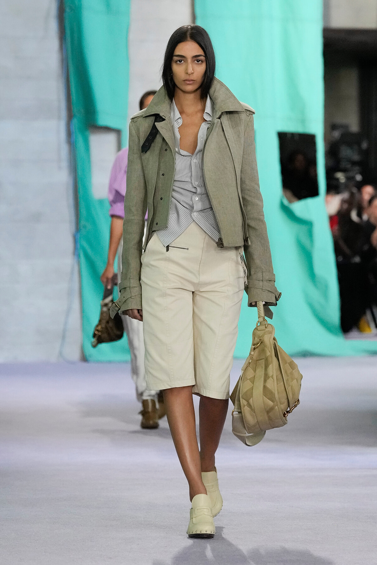 Female model walks a fashion runway in a light gray-green jacket over a striped shirt and beige shorts, carrying a large tan quilted bag.