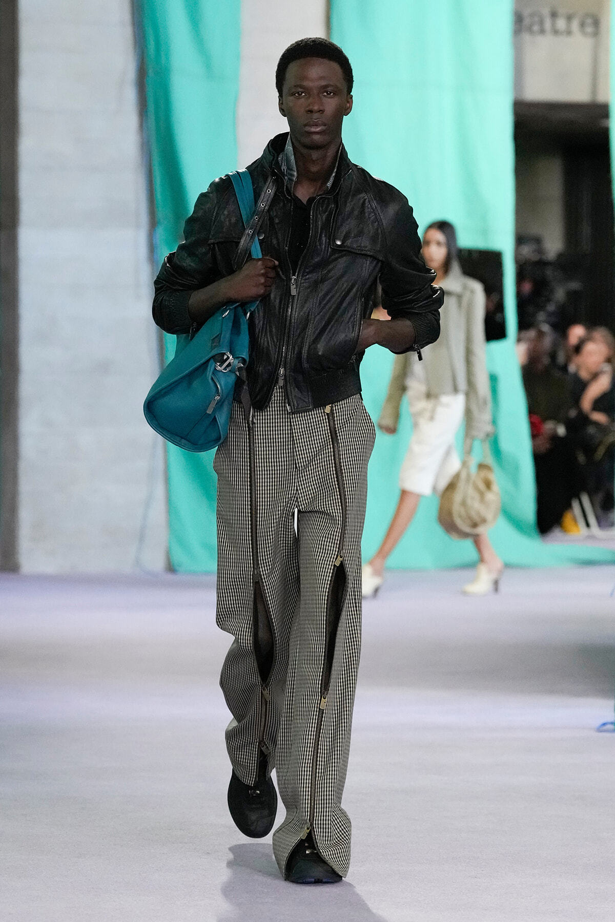 Male fashion model walking a runway with a teal shoulder bag, wearing a black leather jacket and striped wide-leg pants.