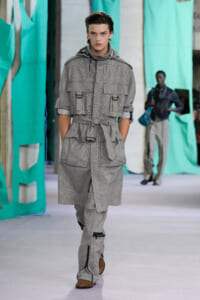 Male model walks a runway in a gray plaid utility jumpsuit with a belted waist and large pockets.
