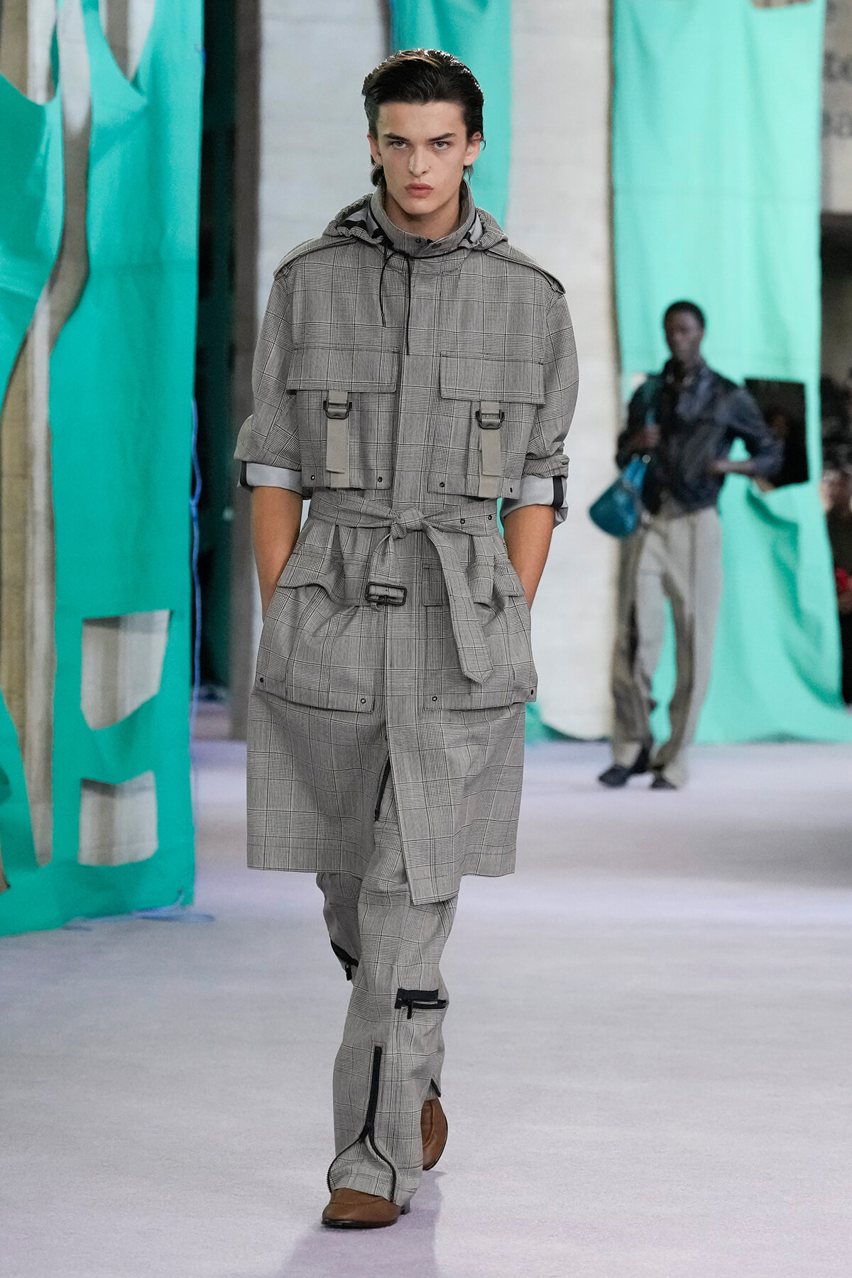 Male model walks a runway in a gray plaid utility jumpsuit with a belted waist and large pockets.