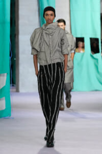 Female model walks a runway wearing a gray striped blouse with a large bow and black-and-white pinstriped pants, head covered with a black cap.