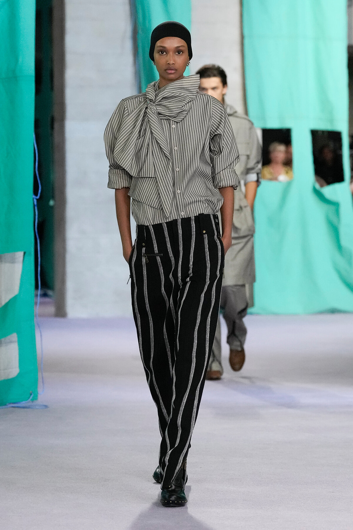 Female model walks a runway wearing a gray striped blouse with a large bow and black-and-white pinstriped pants, head covered with a black cap.