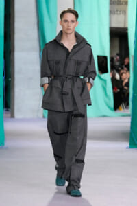 Male model walks the runway in a dark gray utility jumpsuit with multiple pockets and a belt.”,