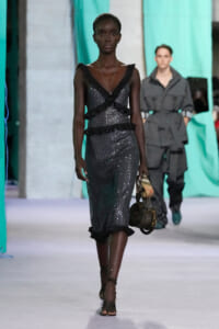 Model walking a runway in a dark sequined dress with black ruffle layers, carrying a small handbag and scarf detail.