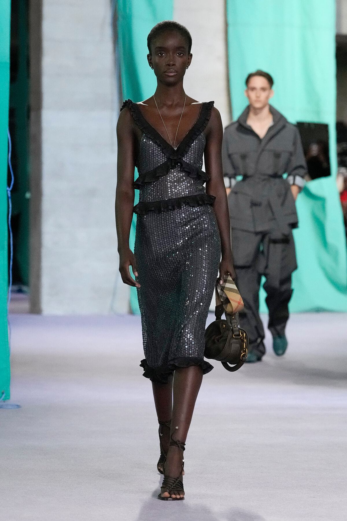 Model walking a runway in a dark sequined dress with black ruffle layers, carrying a small handbag and scarf detail.