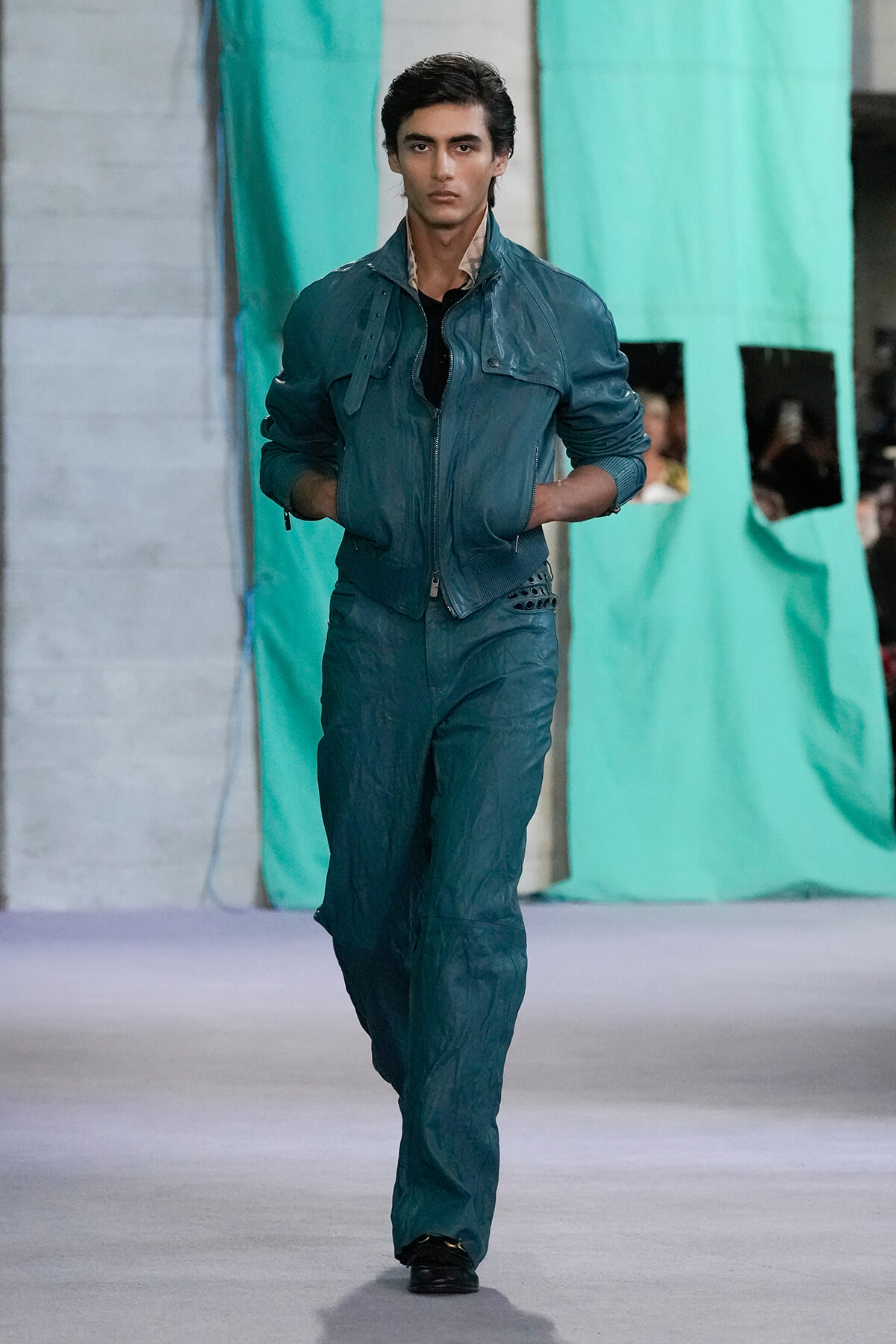 Male model walks the runway in a teal zip-front jacket and matching pants, hands in pockets, turquoise backdrop behind him.