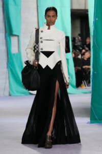 Fashion model on a runway wearing a sculptural white blazer with black accents, a sheer black skirt, and a black handbag.