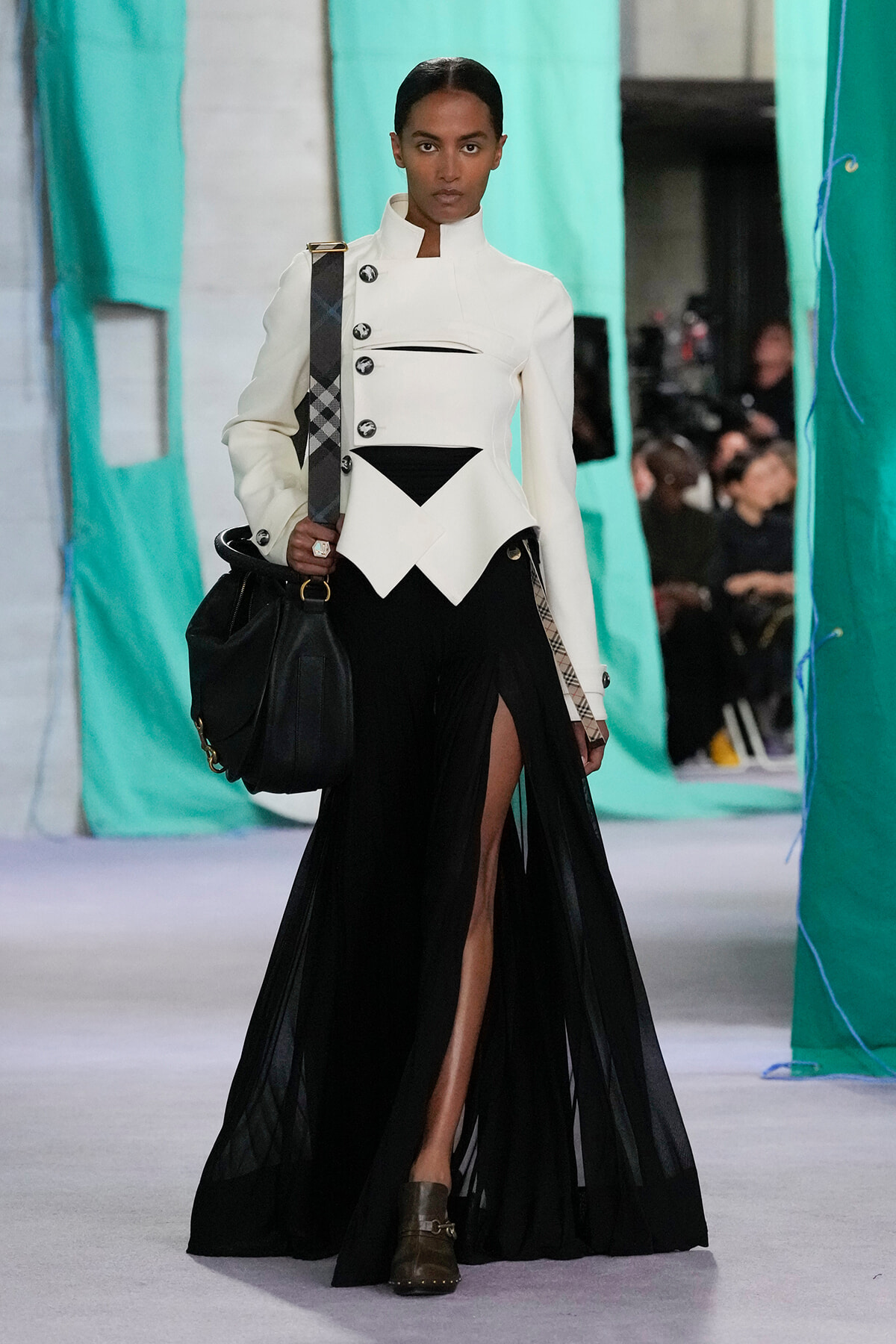 Fashion model on a runway wearing a sculptural white blazer with black accents, a sheer black skirt, and a black handbag.