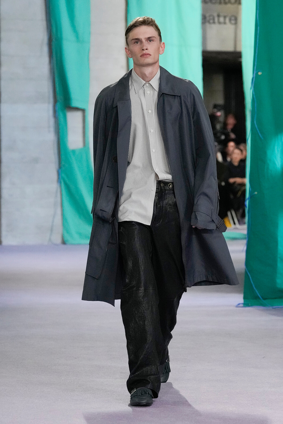 Male model walks a runway in a dark trench coat over a light striped button-down shirt and black pants.