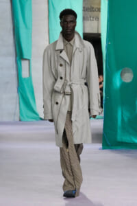 Male model walking the runway in a light gray belted trench coat over a beige shirt and checkered pants.