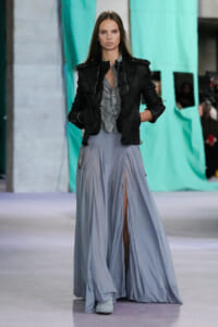 Model walking a runway in a black leather jacket over a gray striped blouse and a flowing pale blue maxi skirt with a front slit.