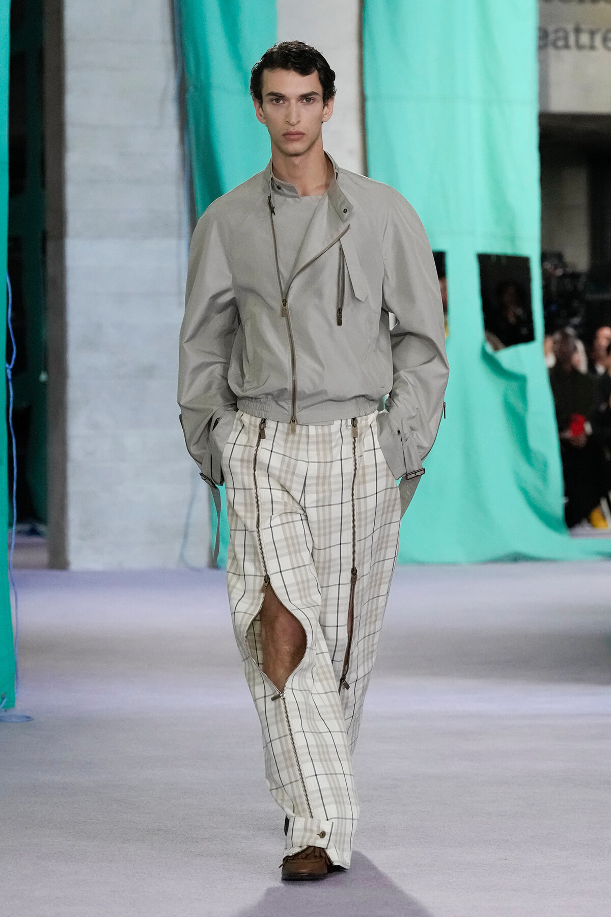 Male fashion model walking a runway in a light gray zip-front jacket and white plaid pants with zipper details.