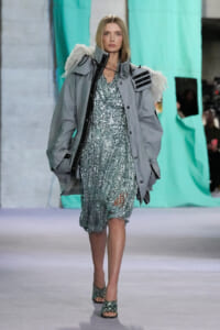 Model walks the runway in a metallic silver-sequin dress under a oversized gray parka with fur-trimmed shoulders during a fashion show