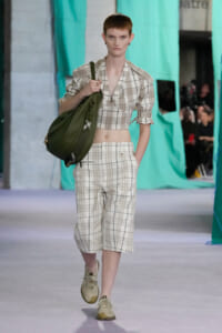 Model walking a fashion show in a beige plaid cropped jacket and matching plaid culottes, carrying an olive green shoulder bag.