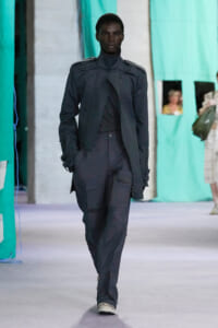 Male model walking a runway in a dark tactical jacket and cargo pants with a serious expression.
