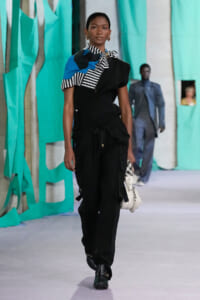 Model walking a runway in a black utility outfit with a striped black-and-white scarf and blue accents, turquoise backdrop visible behind her.