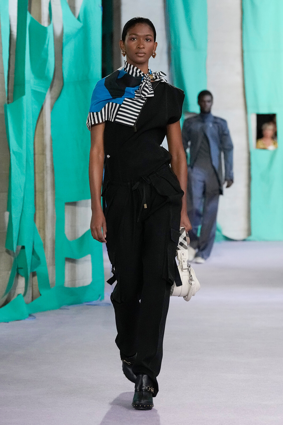 Model walking a runway in a black utility outfit with a striped black-and-white scarf and blue accents, turquoise backdrop visible behind her.
