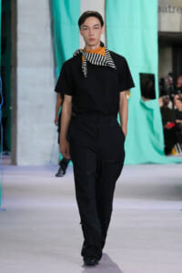 Male model walking runway in black t-shirt and cargo-style black pants, orange top visible under scarf with black-and-white stripes.