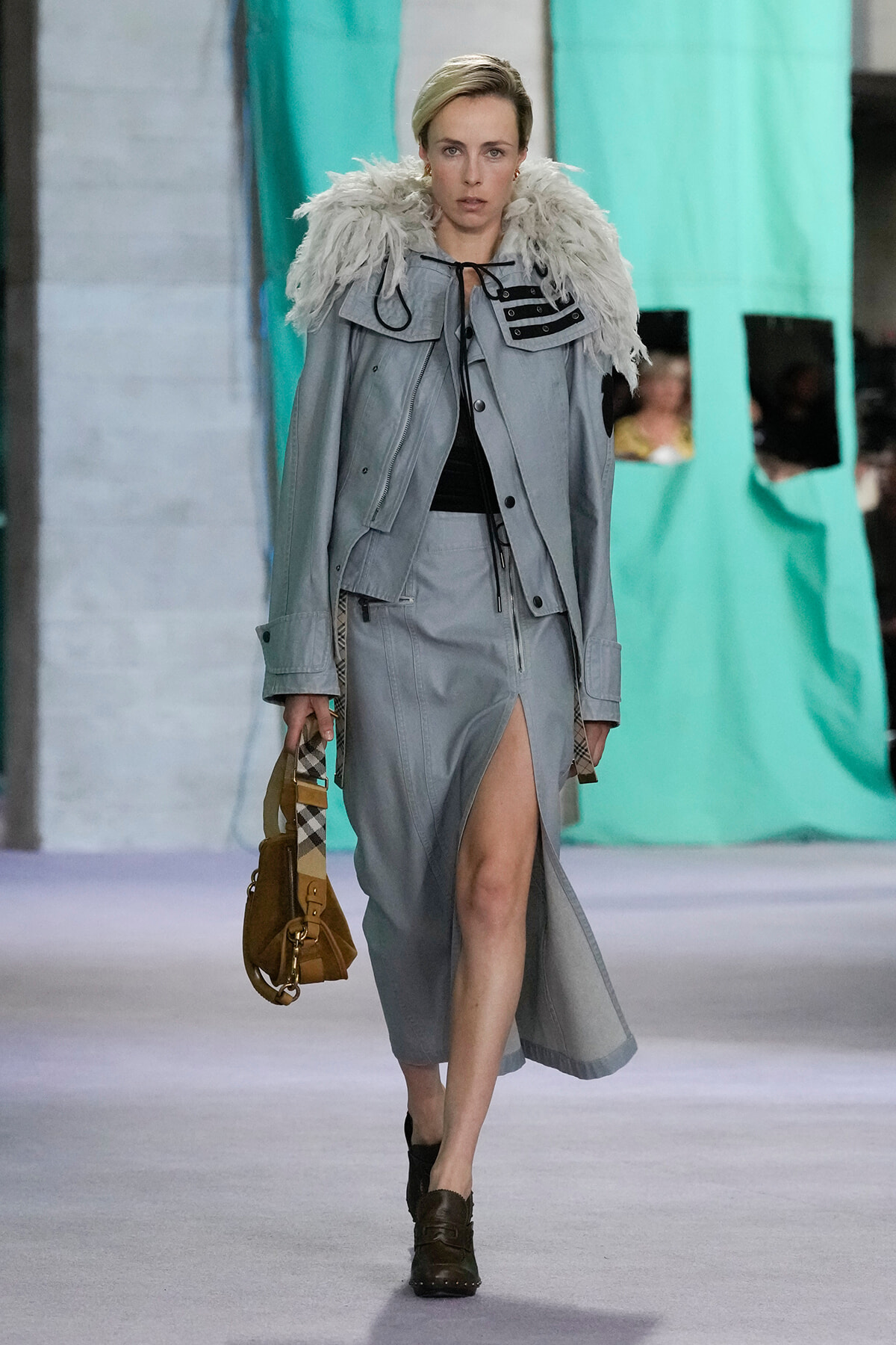 Model walks a runway in a light gray coat with a fluffy feathered collar, matching gray skirt with a front slit, carrying a tan handbag with a plaid strap.