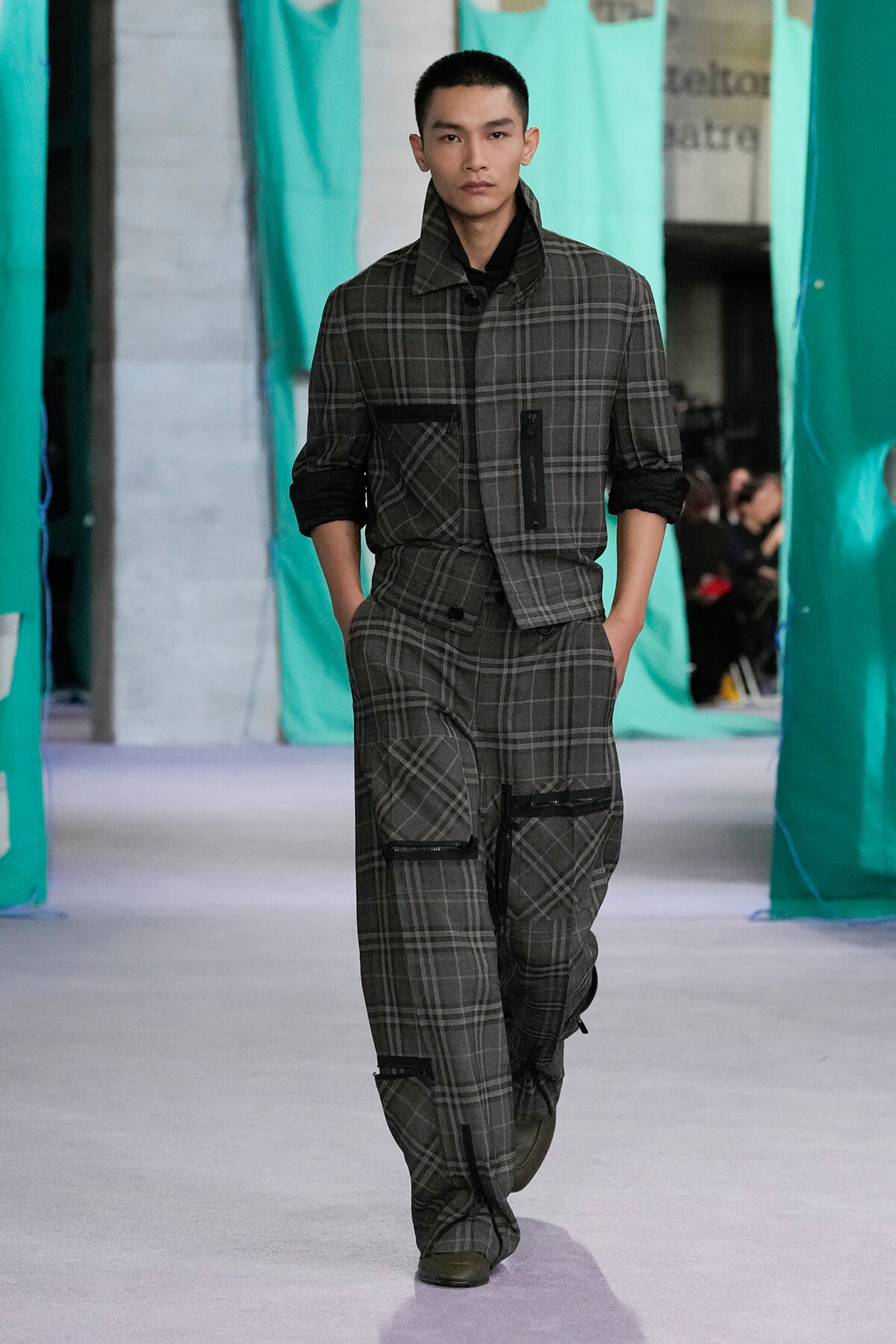 Male model walking a runway in a gray plaid suit with multiple zippered pockets and rolled sleeves.