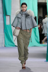 Model walks the runway in a light gray cape with fur trim, loose olive-green pants, and a small white shoulder bag.