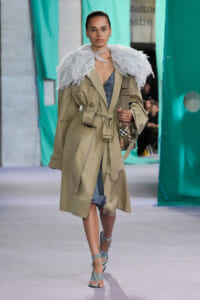 Model walks the runway in a beige trench coat with a fluffy light-gray feather cape, blue inner garment, and strappy sandals with a plaid handbag.
