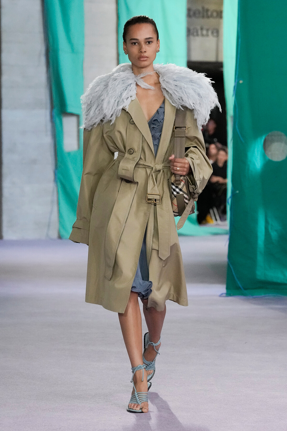 Model walks the runway in a beige trench coat with a fluffy light-gray feather cape, blue inner garment, and strappy sandals with a plaid handbag.
