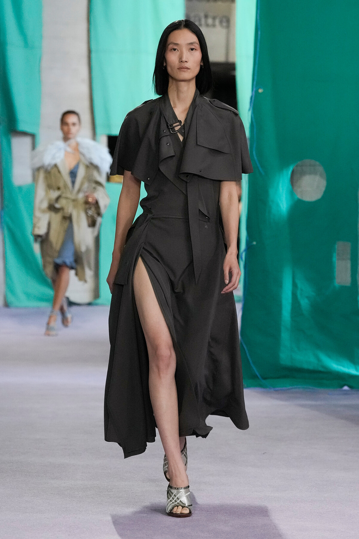 Model walking a dark, oversized trench-style dress with a high slit down the leg on a runway with teal backdrops behind her