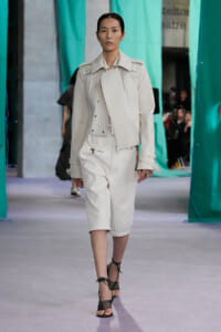 Model walking the runway in an all-white ensemble featuring a structured jacket and knee-length shorts, paired with black strappy heels.