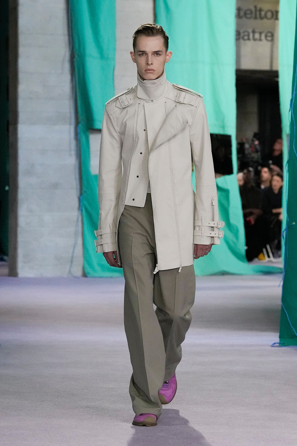 Male model walks a fashion runway in a white, structured asymmetric trench with buckled cuffs and taupe wide-leg pants; pink shoes stand out.