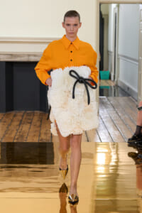 Model walking a fashion runway in an orange shirt and cream textured skirt with a black ribbon belt, wearing gold pointed-toe flats.