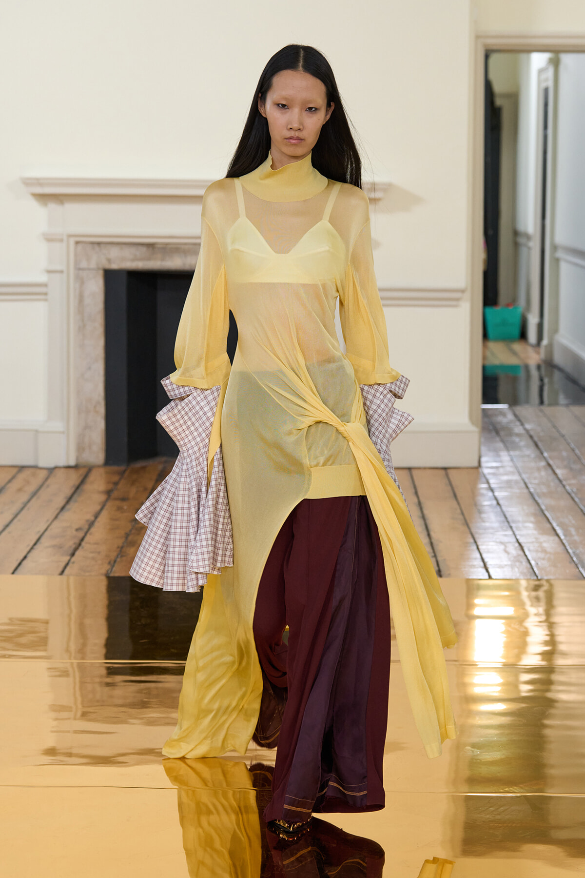 Runway model in a pale yellow sheer dress over a turtleneck, with burgundy pants and plaid-ruffled sleeves, walking a glossy gold runway.