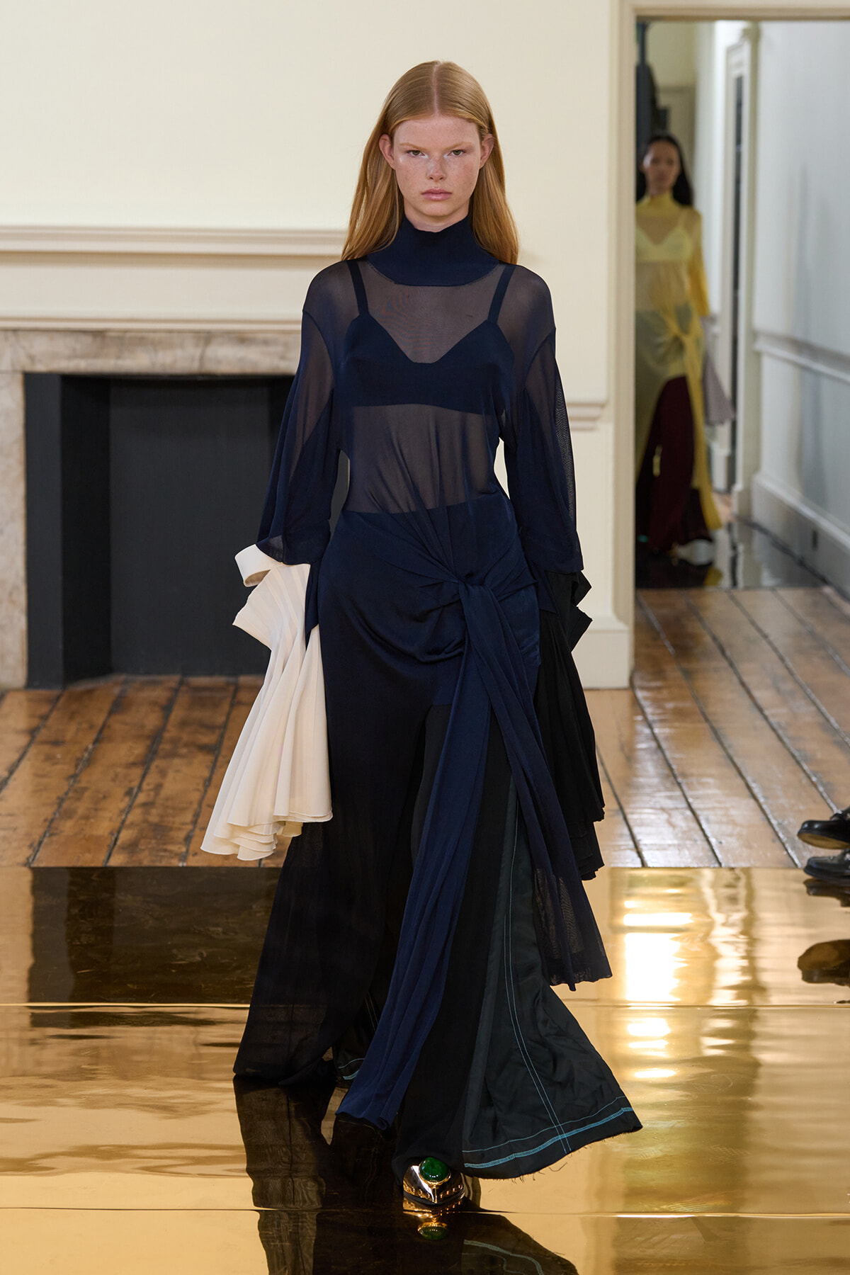 Model walking a fashion show in a navy sheer top over a dark layered dress, with a large fabric knot at the waist and a white draped accessory on her left arm.