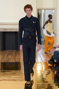 Male model in a navy suit with a black shirt and gold pointed shoes walking the runway.