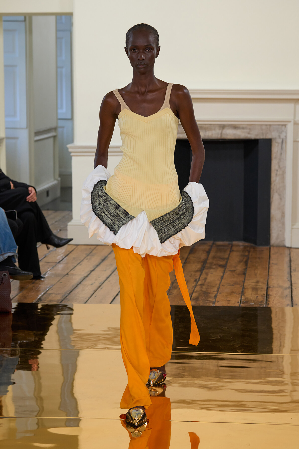 Dark-skinned model walking a fashion runway in a pale yellow ribbed tank top and bright orange pants, with white and gray ruffled fabric draped around the hips.