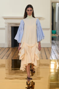 Female model walking the runway in a cream dress with pale blue puff sleeves and a layered ruffled skirt.