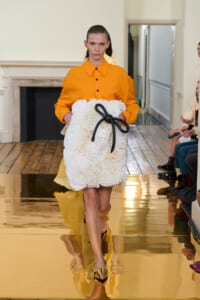 Model on a runway wearing an orange button-down shirt and a fluffy white textured clutch tied with a black bow.",