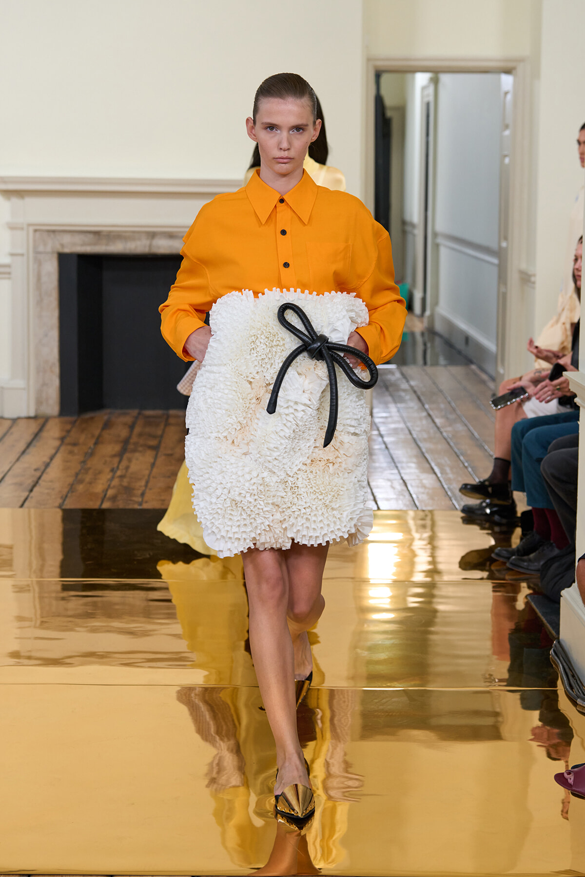 Model on a runway wearing an orange button-down shirt and a fluffy white textured clutch tied with a black bow.",