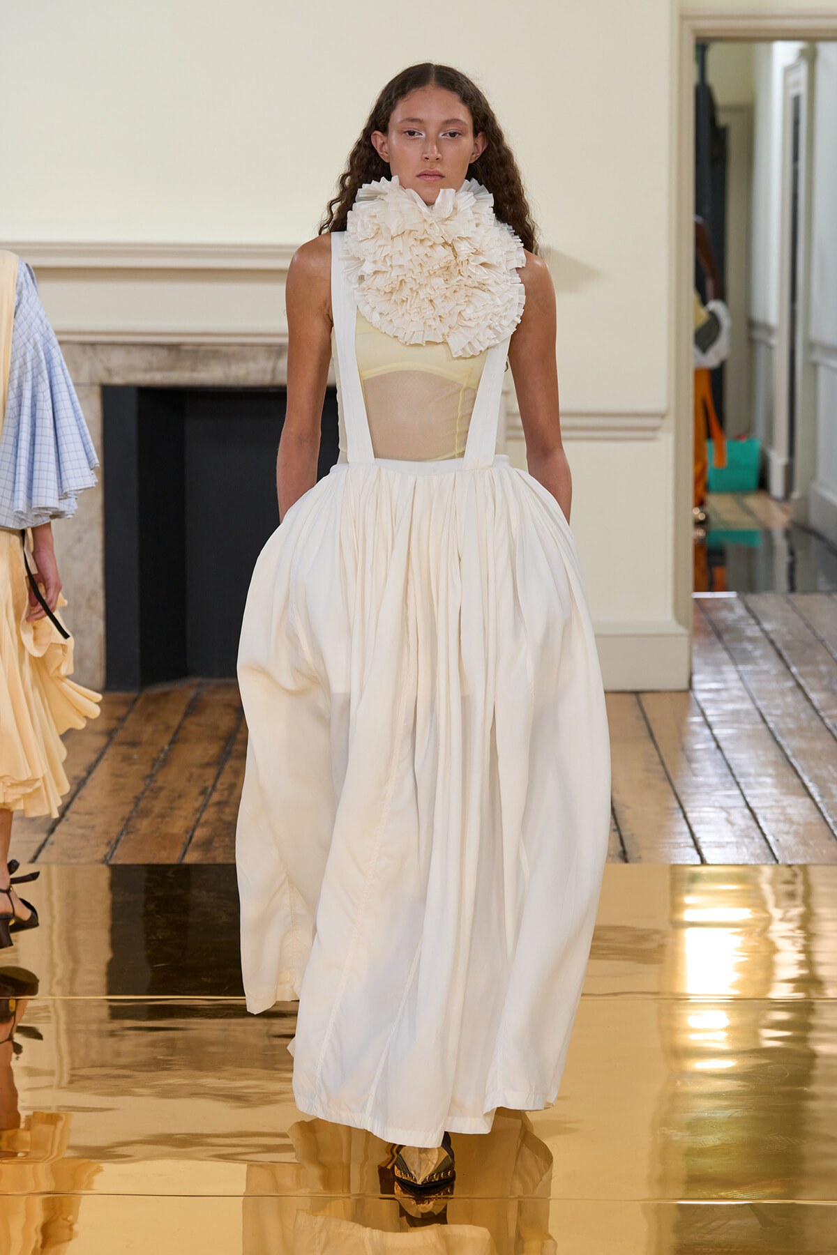 Model walks a runway in an ivory ballgown with a voluminous skirt and a large ruffled collar. The sheer bodice reveals a pale top underneath, set in a neutral backstage-like room with polished floorboards nearby.
