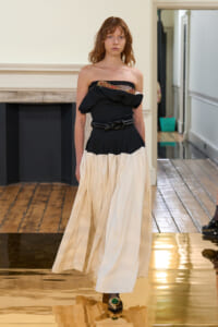 Fashion model walking a runway in a strapless black top with a ruffled edge and floral embellishment, paired with a cream full skirt and a braided black belt on a reflective gold runway.