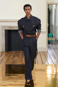 Male model walking a fashion runway in a dark denim jumpsuit with a brown belt and sleeves rolled up; gold shoes visible on a glossy runway surface.