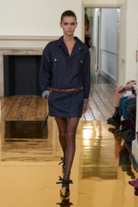 Model walks the runway in a dark denim shirt-dress with a brown belt and rolled cuffs, on a glossy gold floor.