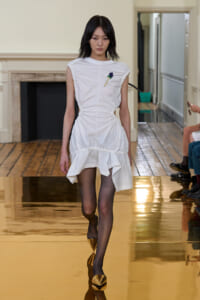 Model walking a white ruched dress with ruffled hem on a gold runway in a fashion show, wearing black tights and gold pointed flats.