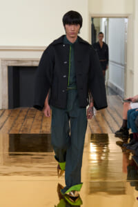 Model walks down a runway in an oversized black coat over green shirt and pants, paired with gold spiked shoes on a reflective gold floor.