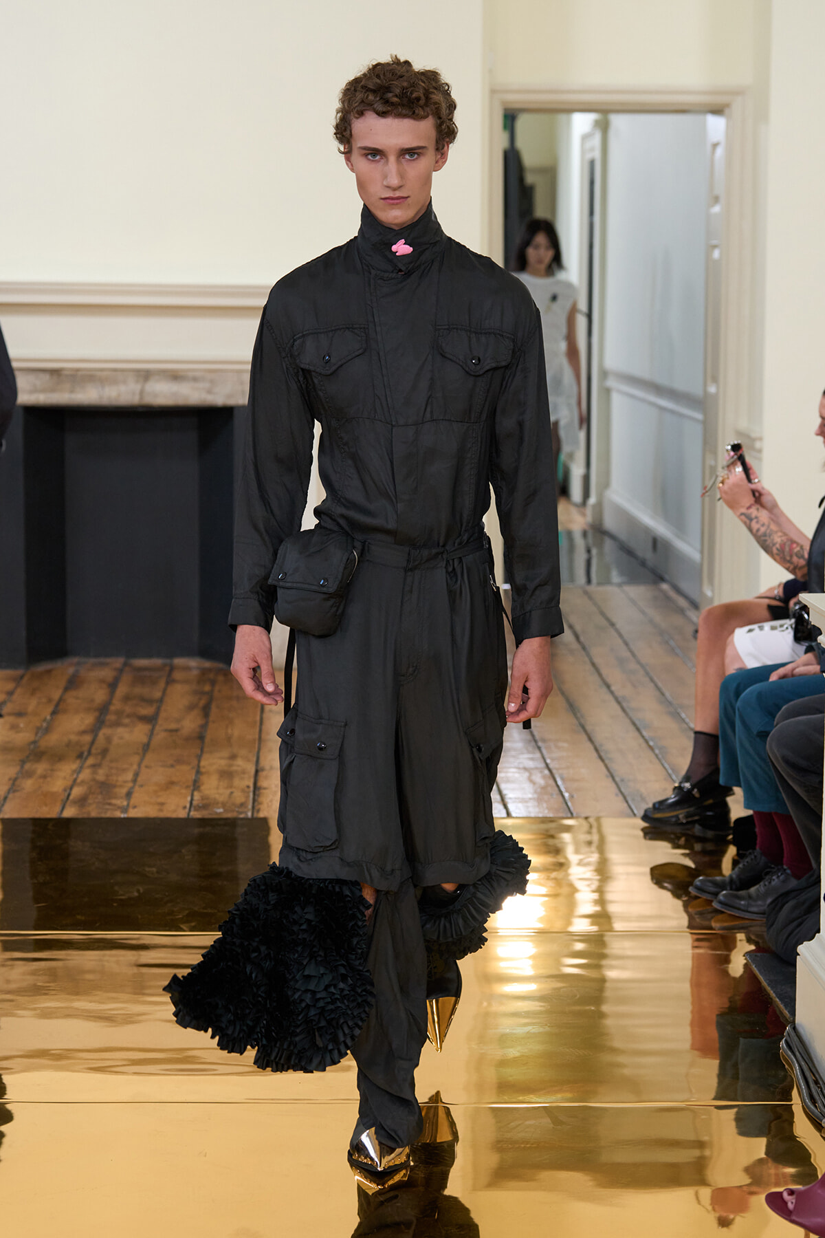 Male model wearing a black utility jumpsuit with a belt bag walking a gold, reflective runway with audience on the sides.
