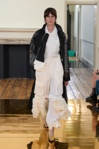 Female runway model wearing white cargo pants with layered ruffles, a white shirt, and a black sheer jacket on a glossy runway.