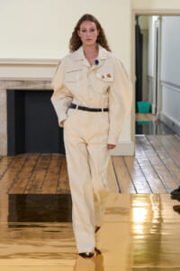 Model walking a runway in a cream utility jumpsuit with oversized sleeves, a black belt, and decorative pins on the chest pockets.
